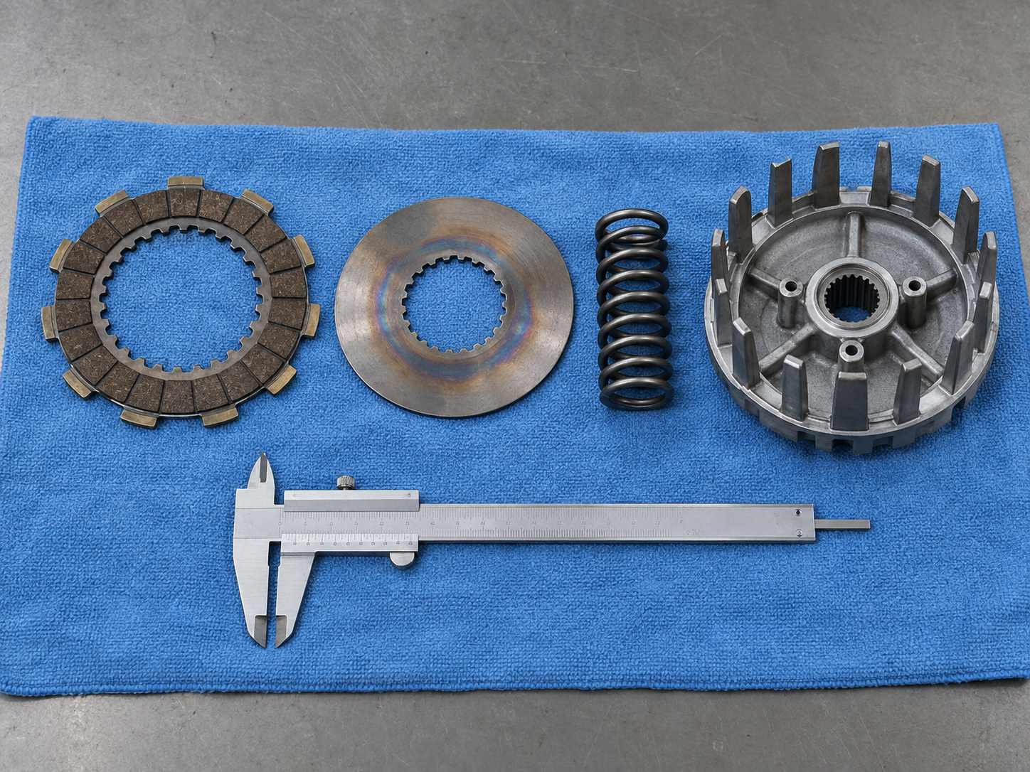 Disassembled pit bike clutch pack components including a friction plate, steel plate, spring, and clutch basket arranged on a workbench for inspection.